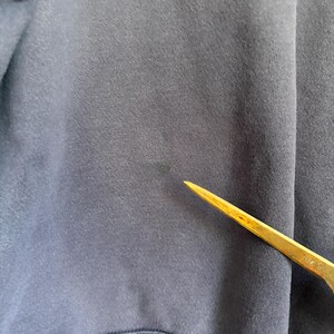 May include: A dark navy blue fabric garment featuring a visible, slightly faded circular stain on the surface. A thin, pointed, metallic gold-colored tool is positioned diagonally across the lower right section of the fabric, pointing toward the stain.