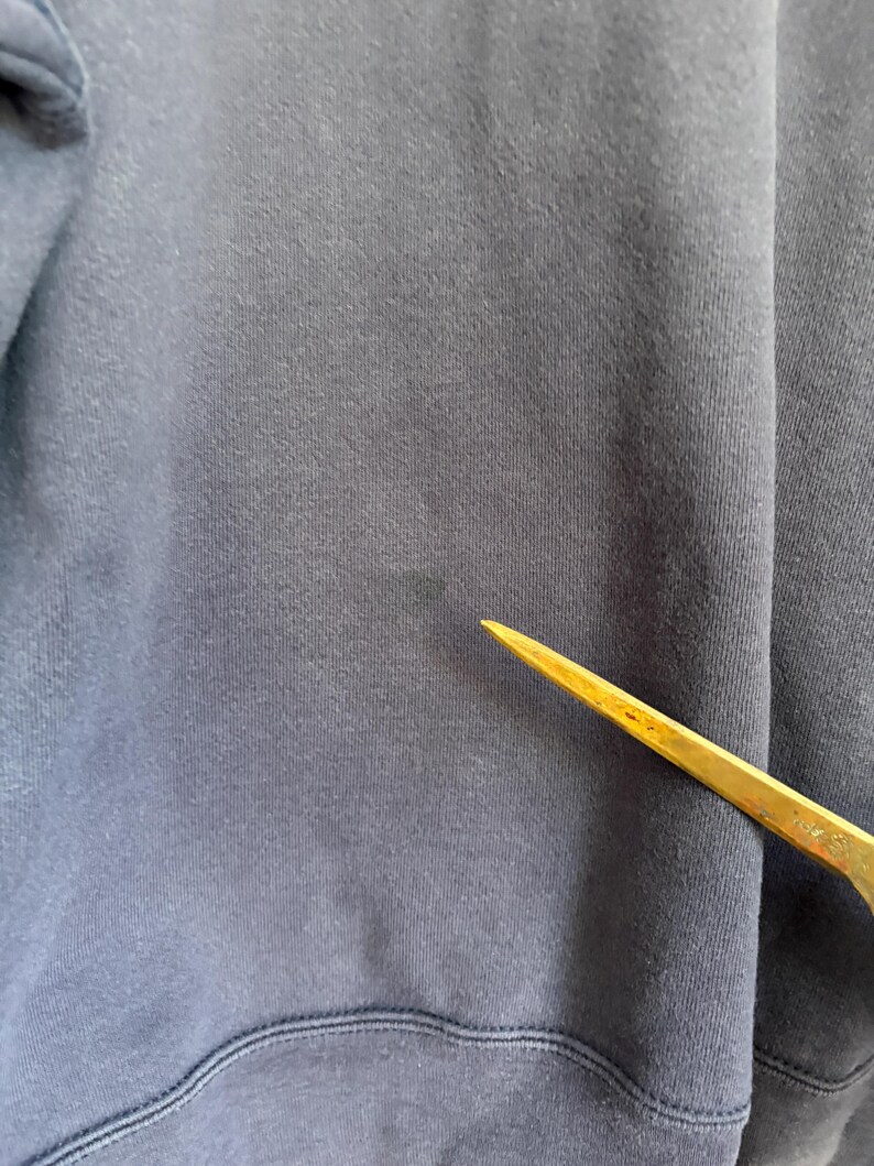 May include: A dark navy blue fabric garment featuring a visible, slightly faded circular stain on the surface. A thin, pointed, metallic gold-colored tool is positioned diagonally across the lower right section of the fabric, pointing toward the stain.