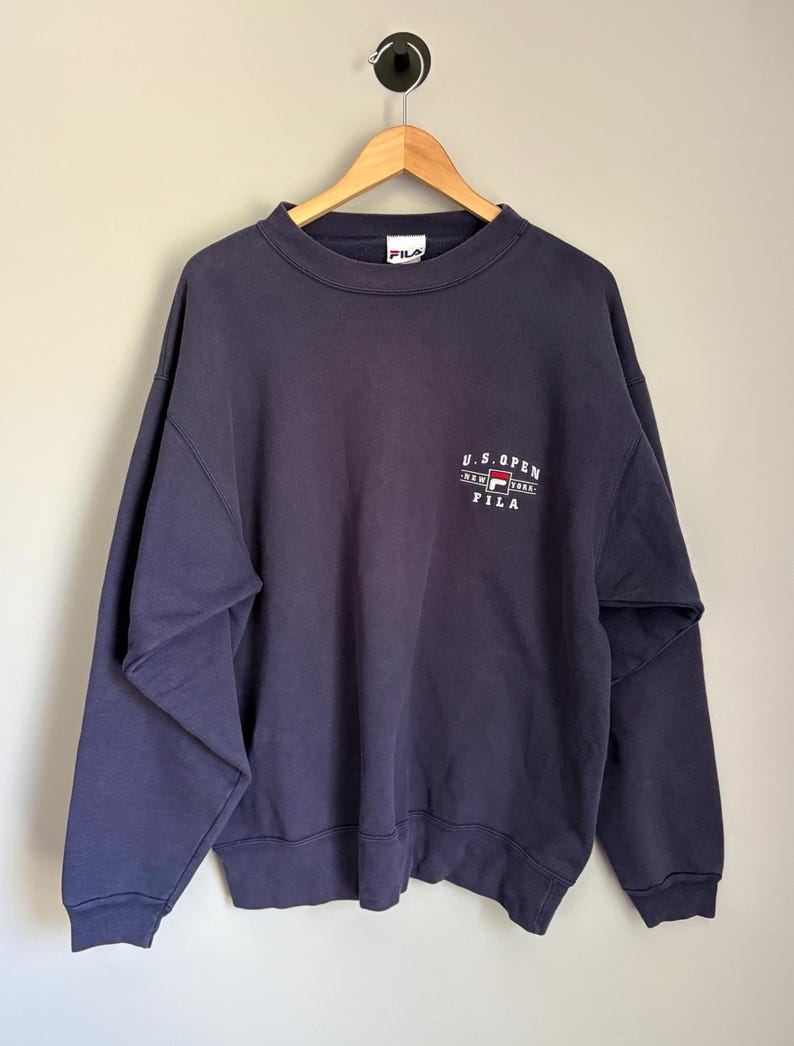 May include: A navy blue long-sleeve crewneck sweatshirt featuring the embroidered text U.S. OPEN NEW YORK FILA on the upper chest. The garment has a ribbed collar, cuffs, and hem, and includes a FILA brand label inside the neckline.