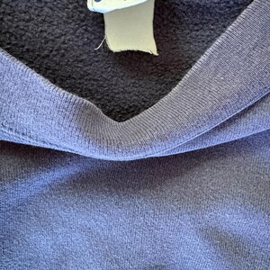 May include: A dark blue sweatshirt featuring a ribbed crew neckline and a white fabric tag sewn into the collar. The tag displays the Fila brand name in blue letters with a red accent mark above the letter i, along with a registered trademark symbol.