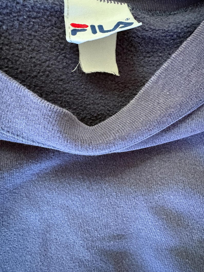 May include: A dark blue sweatshirt featuring a ribbed crew neckline and a white fabric tag sewn into the collar. The tag displays the Fila brand name in blue letters with a red accent mark above the letter i, along with a registered trademark symbol.