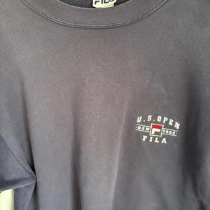 May include: A navy blue crewneck sweatshirt featuring a white and red Fila logo on the chest with the text U.S. OPEN NEW YORK FILA. The garment has a ribbed collar and a white interior neck label with the blue and red Fila brand logo printed on it.