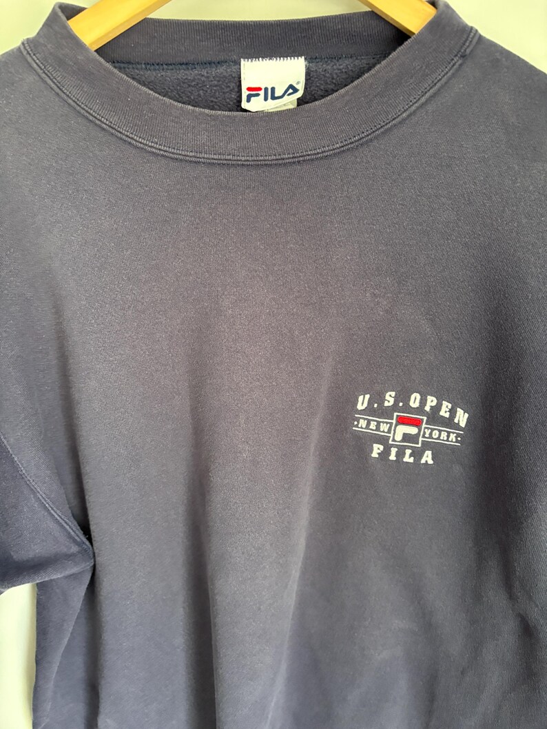 May include: A navy blue crewneck sweatshirt featuring a white and red Fila logo on the chest with the text U.S. OPEN NEW YORK FILA. The garment has a ribbed collar and a white interior neck label with the blue and red Fila brand logo printed on it.