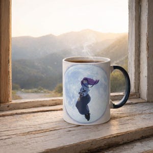 May include: A white ceramic mug with a dark handle featuring a graphic of a person with long purple hair floating in front of a large, pale blue full moon. The mug contains a dark liquid and is positioned on a wooden surface near a window.