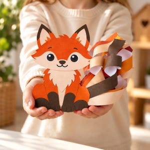 May include: A paper craft fox figure featuring orange, white, and dark brown cardstock layers. The fox has a smiling face, pointed ears, and a large, bushy tail constructed from curled paper strips in shades of orange, brown, and cream.
