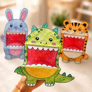 Pode incluir: Três figuras de papel em forma de coelho azul, dinossauro verde e tigre laranja. Cada figura apresenta uma boca larga e aberta com coloração vermelha no interior e dentes brancos, concebida como uma personagem dobrável com base plana.
