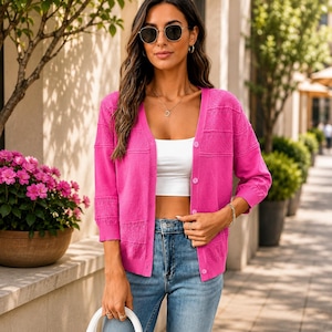 May include: A bright pink button-down cardigan featuring three-quarter length sleeves and a textured knit pattern. The garment is worn over a plain white crop top and blue denim jeans, showcasing a V-neckline and a series of matching pink buttons.