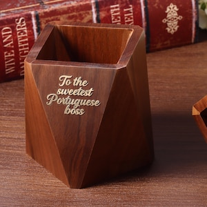 Könnte beinhalten: Ein facettierter Stiftehalter aus Holz mit dunkelbrauner Maserung. Auf der Vorderseite ist der Text "To the sweetest Portuguese boss" in einer hellen Schreibschrift eingraviert. Der Behälter hat eine geometrische Form mit scharfen Kanten.