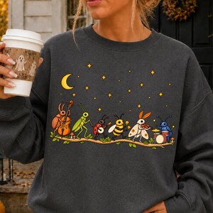 Puede incluir: Una sudadera gris oscuro de cuello redondo con un gráfico de varios insectos tocando instrumentos musicales sobre una rama bajo una luna creciente y estrellas. Los insectos incluyen un escarabajo, un saltamontes, una mariquita, una abeja y una polilla.