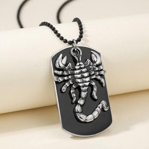 May include: A silver-toned metal dog tag pendant featuring a raised, detailed scorpion figure centered on a smooth, glossy black rectangular background. The pendant hangs from a thin black beaded chain, creating a high-contrast accessory piece.