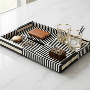May include: A rectangular tray featuring a black and white striped pattern with a gold-toned nameplate. The tray holds a tan leather wallet, a black pen, a gold pocket watch with a chain, a small wooden box, and two clear glasses with gold rims.