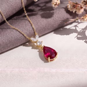 Puede incluir: Un collar de oro con un colgante de piedra preciosa roja en forma de pera suspendido de una cadena delicada. El colgante está adornado con dos pequeñas piedras claras de talla marquesa dispuestas en forma de hoja sobre la piedra roja.