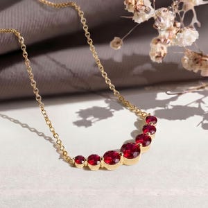May include: A gold chain necklace featuring a graduated row of six round, faceted red gemstones set in gold bezels. The stones increase in size toward the center, creating a curved, symmetrical arrangement along the delicate metal link chain.