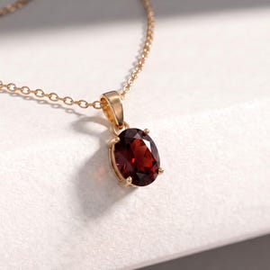 May include: A gold necklace featuring an oval-cut deep red garnet gemstone pendant. The faceted stone is held in a four-prong setting attached to a delicate gold chain. The pendant hangs from a simple gold bail, showcasing the dark red color.