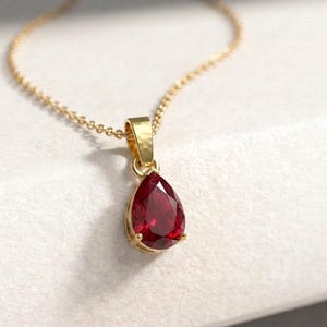 May include: A gold necklace featuring a pear-shaped, deep red gemstone pendant. The faceted ruby-coloured stone is set in a polished gold frame with a small bail, suspended from a delicate gold chain that curves gracefully across the frame.
