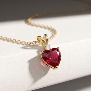 May include: A gold necklace featuring a heart-shaped red gemstone pendant. The pendant is suspended from a delicate gold chain and is accented by a single small, clear round gemstone set into the gold bail above the heart-shaped stone.