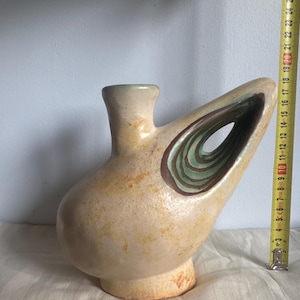 Może przedstawiać: Ceramiczny wazon z kremowo-brązowym nakrapianym szkliwem, posiadający centralną szyjkę oraz duży, wydrążony uchwyt boczny z detalami w postaci zielonych i brązowych koncentrycznych pierścieni. Żółta miarka wskazuje wysokość około 24 cm.