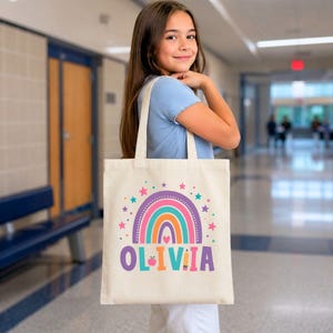May include: A cream-colored canvas tote bag featuring a colorful rainbow graphic with stars and hearts. The name OLIVIA is printed in large, multi-colored letters at the bottom, with a small apple icon replacing the letter I and a pencil icon.