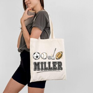 May include: A cream-colored canvas tote bag featuring a graphic print of a soccer ball, baseball, golf club with ball, and an American football. Below the sports equipment, the name MILLER is printed in a bold, black, block-style font.