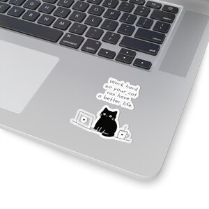 Könnte beinhalten: Ein gestanzter Aufkleber mit einer schwarzen Katze, die vor einem Laptop neben einer Kaffeetasse sitzt. Über der Katze steht der Text: Work hard so your cat can have a better life. Der Aufkleber befindet sich auf der Oberfläche eines silbernen Laptops.
