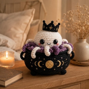 May include: A crocheted white octopus with black eyes and pink cheeks wearing a black crown, sitting inside a black crocheted cauldron. The cauldron features gold embroidered moon phases and stars, with purple and white tentacles spilling over the rim.