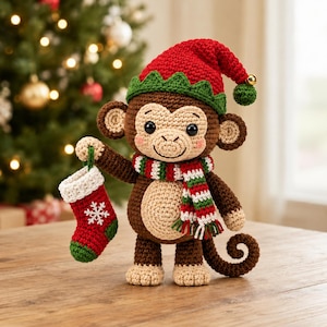 Puede incluir: Un mono de ganchillo marrón y beige que lleva un gorro de elfo rojo y verde con un pequeño cascabel dorado y una bufanda a rayas roja, blanca y verde. El mono sostiene un pequeño calcetín navideño de ganchillo rojo y verde con un copo.