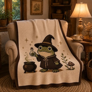May include: A cream-colored knit throw blanket featuring a dark brown border and a central graphic of a green frog wearing a pointed wizard hat and robe. The frog holds a potion bottle near a black cauldron with green smoke, surrounded by stars.