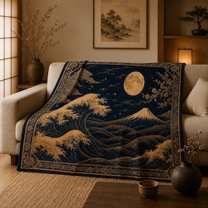 May include: A woven throw blanket featuring a dark blue background with a gold-toned graphic design of a large ocean wave, a mountain peak, a full moon, and starry skies, finished with a decorative floral border pattern along the edges.