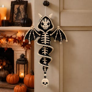 May include: A hanging crochet decoration featuring a black and cream-colored skeletal dragon with wings, a hooded skull head, and a spiraling body. A small cream-colored skull hangs from the bottom, suspended by black and cream beads.