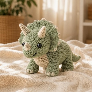 Puede incluir: Un juguete de dinosaurio triceratops de ganchillo hecho con hilo verde salvia. La criatura presenta una gran gola, tres cuernos de color crema, una barriga de color crema y ojos de plástico negro. La textura es consistente en todo el cuerpo.