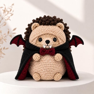 May include: A crocheted hedgehog plush toy featuring a dark brown textured back, a tan face and body, and black plastic eyes. The hedgehog wears a black cape with a deep red lining, small red bat wings, a dark red bow tie, and two white fangs.