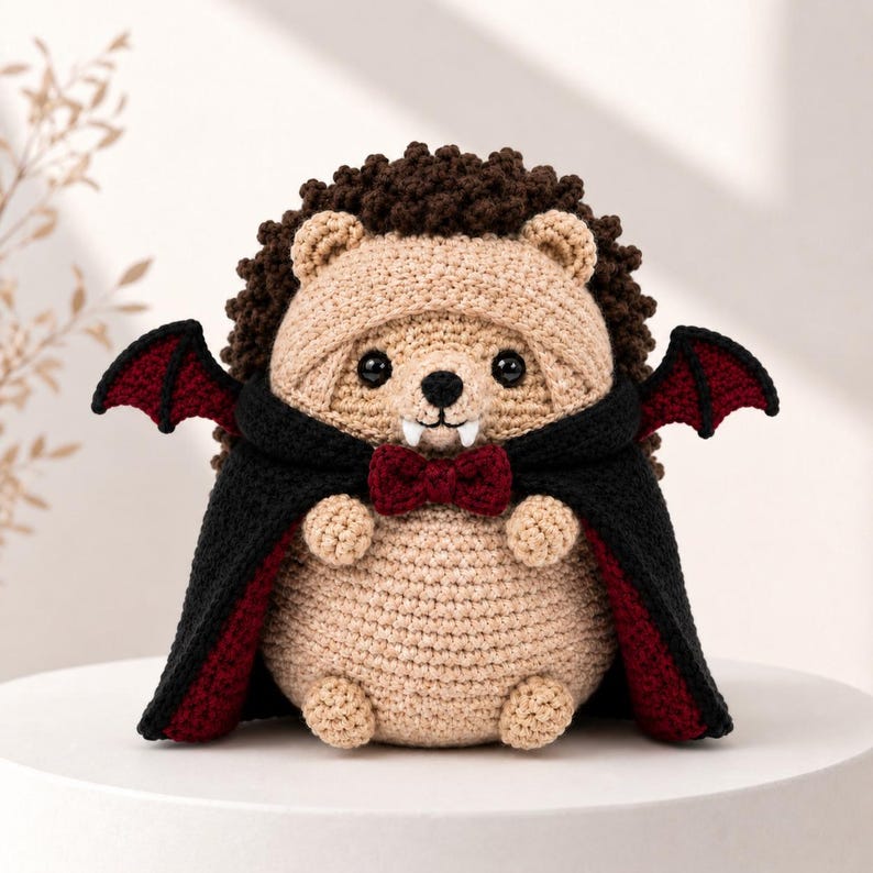 May include: A crocheted hedgehog plush toy featuring a dark brown textured back, a tan face and body, and black plastic eyes. The hedgehog wears a black cape with a deep red lining, small red bat wings, a dark red bow tie, and two white fangs.
