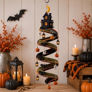 May include: A spiral hanging decoration featuring a crocheted black and green structure topped with a yellow crescent moon and a dark house. It is adorned with small dangling charms including orange pumpkins, white ghosts, black bats, and lanterns.