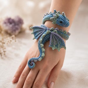 May include: A crochet dragon bracelet featuring a blue and green color palette with purple accents on the wings. The dragon design wraps around the wrist, with the head resting near the forearm and the long, curled tail extending toward the hand.