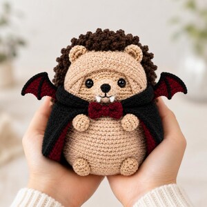 May include: A crocheted hedgehog plush toy featuring a tan body, dark brown textured back, and black vampire cape with a deep red lining. The figure includes small black bat wings, a dark red bow tie, and two white fangs protruding from the mouth.