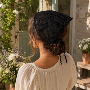 May include: A black triangular headscarf featuring an intricate crocheted spiderweb pattern. The accessory is crafted from fine black thread and includes two thin, curly crocheted ties at the back to secure the head covering in place.