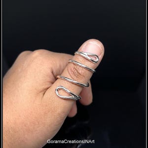 May include: A silver-colored spiral ring that wraps around the finger multiple times. The design features a smooth, polished metallic finish with open, teardrop-shaped ends at the top and bottom of the coil. The text GoramaCreationsINArt is visible.