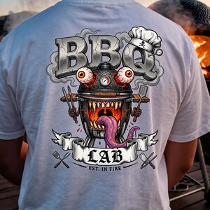 Könnte beinhalten: Ein weißes T-Shirt mit einem Rückenaufdruck eines monströsen Holzkohlegrills mit hervorstehenden Augen, scharfen Zähnen und einer langen Zunge. Das Design enthält den Text BBQ LAB EST. IN FIRE, eine Kochmütze, eine Grillzange, einen Wender und Besteck.
