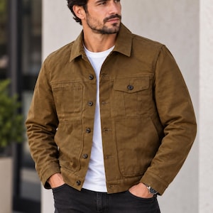 Könnte beinhalten: Eine braune Jacke mit Knopfleiste, spitzem Kragen, langen Ärmeln und zwei Brusttaschen, getragen über einem schlichten weißen T-Shirt und kombiniert mit einer dunkelgrauen Jeans. Die Jacke hat ein strukturiertes Design mit sichtbaren Nähten.