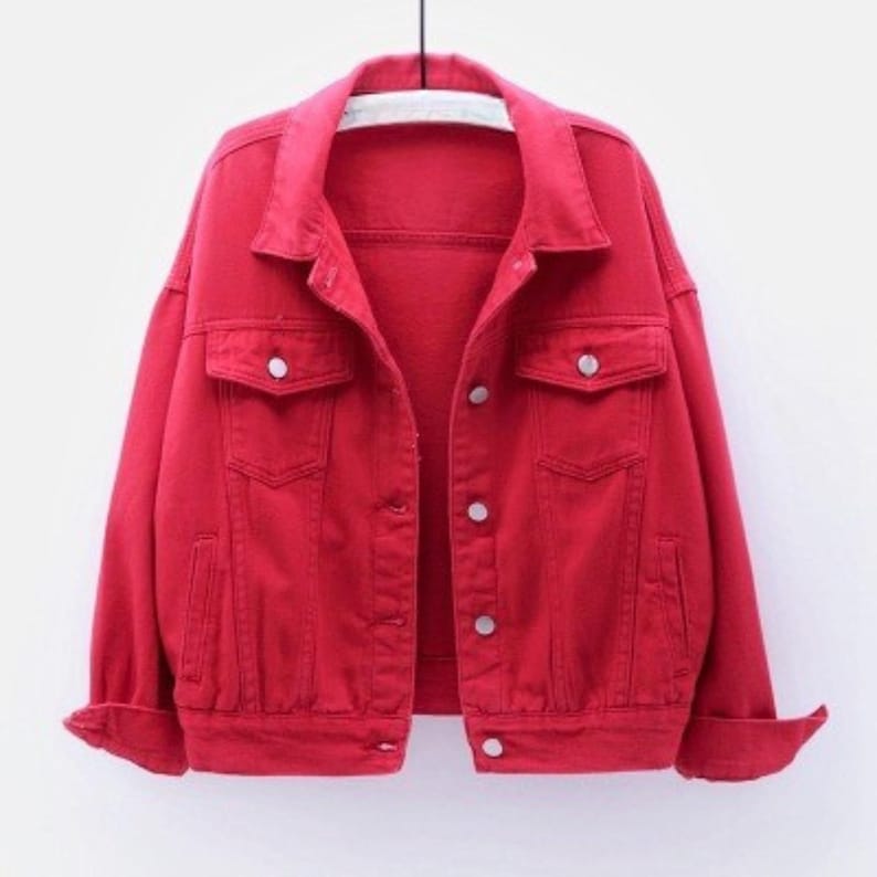 May include: A bright red denim jacket featuring a classic collar, long sleeves with buttoned cuffs, and a front button closure. The jacket includes two chest pockets with buttoned flaps and two side pockets, all constructed from a solid red fabric.