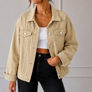 May include: A beige denim jacket featuring a classic collar, long sleeves with cuffs, two chest pockets with button flaps, and a front button closure. The jacket is worn over a white cropped top and paired with black pants, showcasing a structured fit.