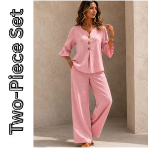 May include: A two-piece set consisting of a light pink top and matching wide-leg trousers. The top features a V-neckline, three-quarter length sleeves with ruffled cuffs, and three large brown buttons down the front. The text Two-Piece Set is visible.