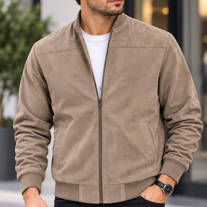 May include: A taupe suede bomber jacket featuring a ribbed collar, cuffs, and hem, a full-length front zipper closure, and horizontal seam detailing across the chest. The jacket is worn over a plain white t-shirt, showcasing a clean design.