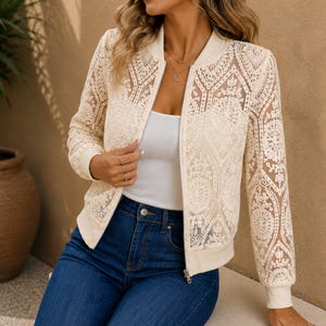 May include: A cream-colored bomber jacket featuring an intricate, sheer lace overlay with a detailed floral and geometric pattern. The jacket has a ribbed collar, cuffs, and hem, a full-length front zipper, and long sleeves, worn over a white top.