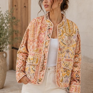 May include: A quilted jacket featuring a vibrant paisley pattern in shades of orange, yellow, pink, and purple. The jacket has a round neckline, long sleeves, and two front patch pockets with flaps, finished with light-colored trim along the edges.