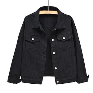 May include: A solid black denim jacket featuring a classic collar, long sleeves with cuffs, and a front button-down closure. The jacket includes two chest pockets with buttoned flaps and visible stitching details throughout the garment structure.