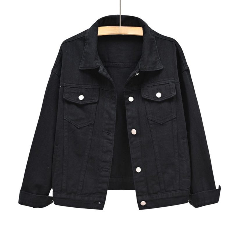 May include: A solid black denim jacket featuring a classic collar, long sleeves with cuffs, and a front button-down closure. The jacket includes two chest pockets with buttoned flaps and visible stitching details throughout the garment structure.