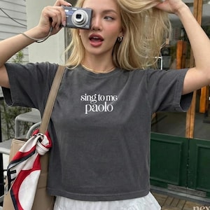 May include: A charcoal gray short-sleeve t-shirt featuring the white text "sing to me paolo" centered on the chest. The shirt has a crew neckline and a relaxed fit, paired with a white skirt and a bag with a red, white, and black patterned scarf.
