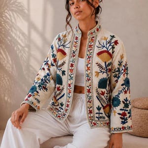 May include: An off-white open-front jacket featuring a mandarin collar and intricate multi-colored floral embroidery in shades of red, blue, and yellow. The garment displays the text ZAHAAR in the upper right corner and has long sleeves.