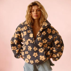 Pode incluir: Um suéter de fleece cinza-carvão com um padrão repetitivo de flores bege claro. A peça possui gola alta, fechamento parcial com zíper na frente, mangas compridas e um bolso canguru grande feito de material texturizado e felpudo.